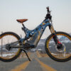 A wide view showing an Optibike E-Bike with snow covered mountains in the background, showing the hand painted frame featuring a wolf howling at the sky, painted in vibrant gold color.