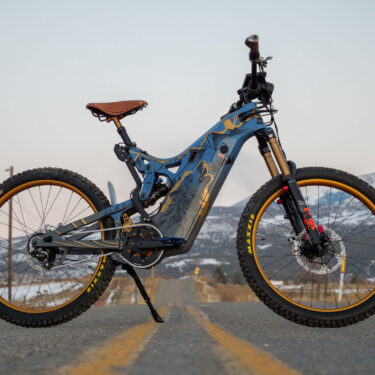 A wide view showing an Optibike E-Bike with snow covered mountains in the background, showing the hand painted frame featuring a wolf howling at the sky, painted in vibrant gold color.