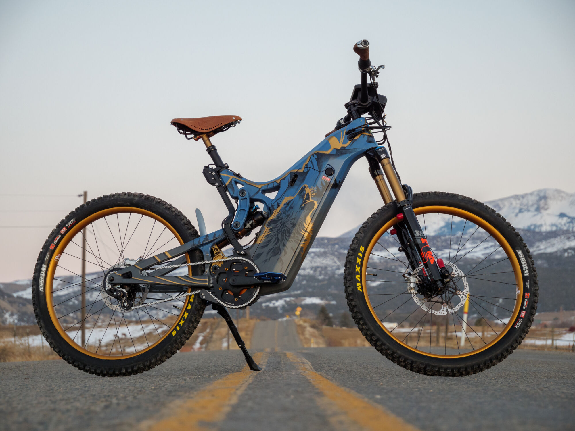 optibike-r17-wolf-paint (2) A wide view showing an Optibike E-Bike with snow covered mountains in the background, showing the hand painted frame featuring a wolf howling at the sky, painted in vibrant gold color.