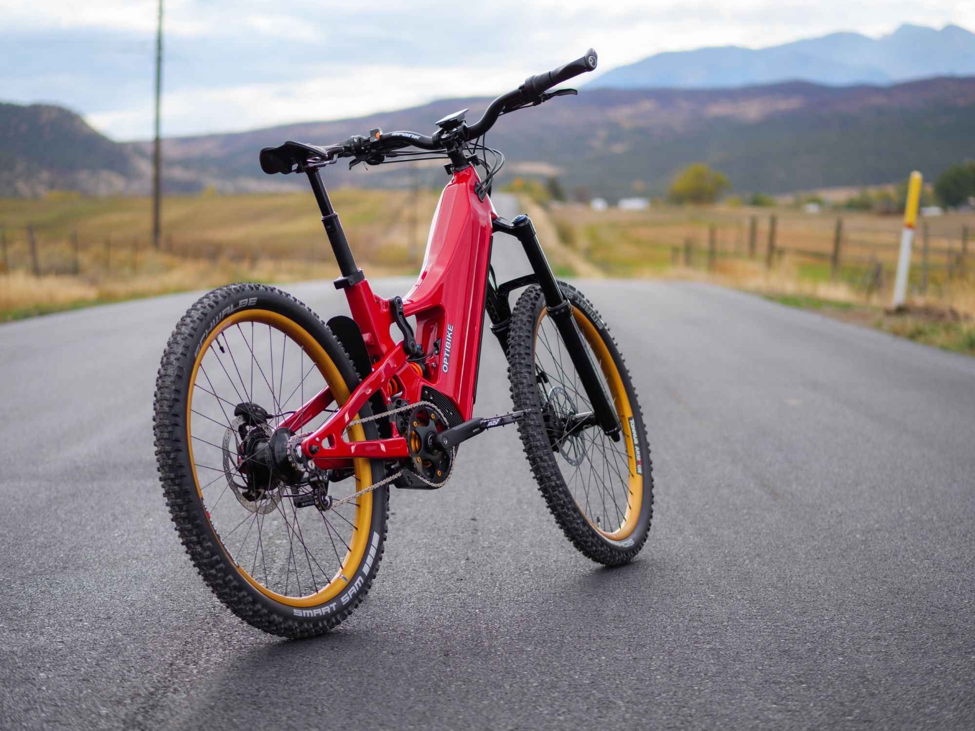 Optibike Loki eMTB angled profile with a red frame, parked on the road with mountains in the background.
